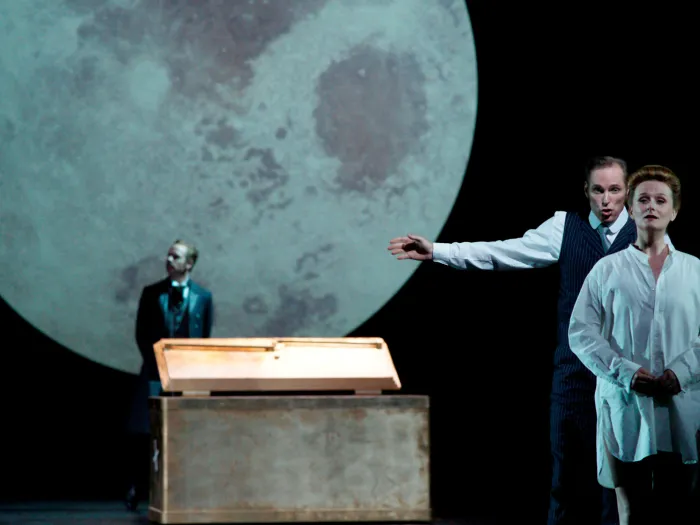 Szene aus "Der Kaufmann von Venedig", 2013 im Festspielhaus, drei kostümierte Darstellende, große Mondprojektion im Hintergrund, zwei Personen rechts im Bild, eine davon mit ausgestrecktem Arm, die dritte Person im Hintergrund