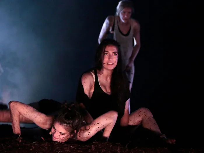 Szene aus "The Wasp Factory", 2013 im Festspielhaus, drei kostümierte Darstellerinnen, eine liegt am Boden mit angewinkelten Armen, eine sitzt dahinter, die dritte steht im Hintergrund, dunkler Bühnenhintergrund mit Nebel