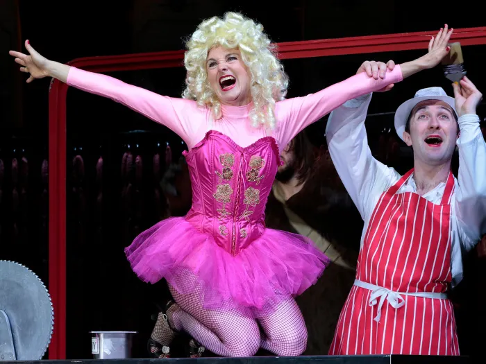 Szene aus "Gloria von Jaxtberg", 2014 im Theater am Kornmarkt, eine Person im pinken Kostüm mit Tutu, blonden Locken und ausgestreckten Armen kniet, rechts hält eine weitere Person einen Arm der knienden Person und trägt einen weißen Hut sowie eine rot-weiß-gestreifte Schürze