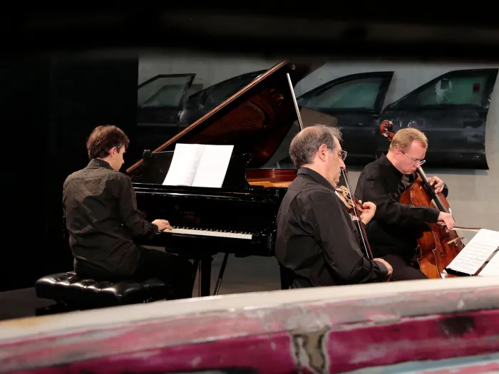 Szene aus "KAZ – Konzerte", 2014 an diversen Spielorten, drei Musiker spielen ihre Instrumente – Klavier, Cello, Violine –, alle in schwarzer Kleidung
