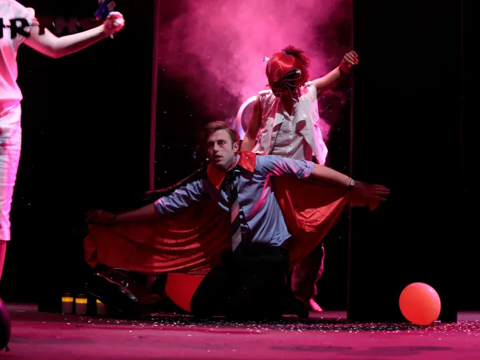 Szene aus "Das Leben am Rande der Milchstraße", 2014 im Festspielhaus, eine Person mit rotem Umhang kniet mit ausgestreckten Armen auf der Bühne, umgeben von anderen Personen, ein roter Luftballon auf dem Boden, rotes Licht