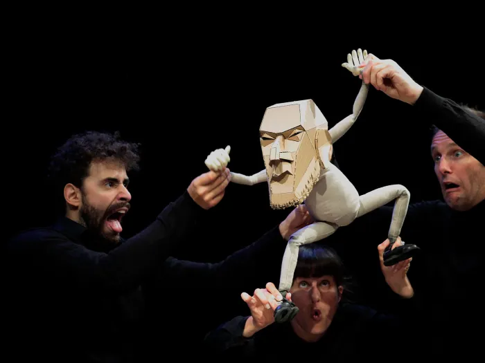 Szene aus "The Table", 2014 im Theater Kosmos, drei Personen in schwarzer Kleidung halten eine weiße Puppe in die Luft