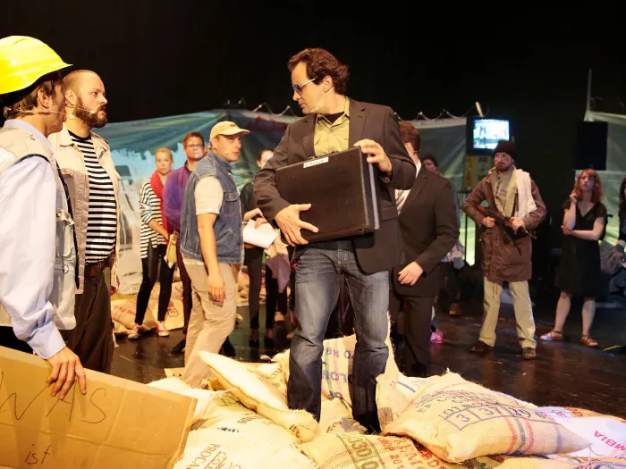 Szene aus "Trans-Maghreb", 2014 im Festspielhaus, drei Personen im Vordergrund – links eine Person mit gelbem Helm und einem Pappkarton in der Hand, mittig ein Mann mit Koffer –, im Hintergrund mehrere Personen