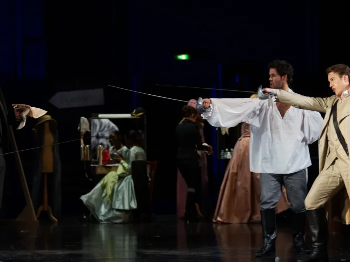 Szene aus "Così fan tutte", 2015 im Theater am Kornmarkt, drei Darsteller im Vordergrund, zwei mit je einem Degen, einer in weißem Hemd und schwarzen Stiefeln, der andere in hellem Mantel, der dritte steht links im schwarzen Anzug, im Hintergrund weitere kostümierte Darstellende