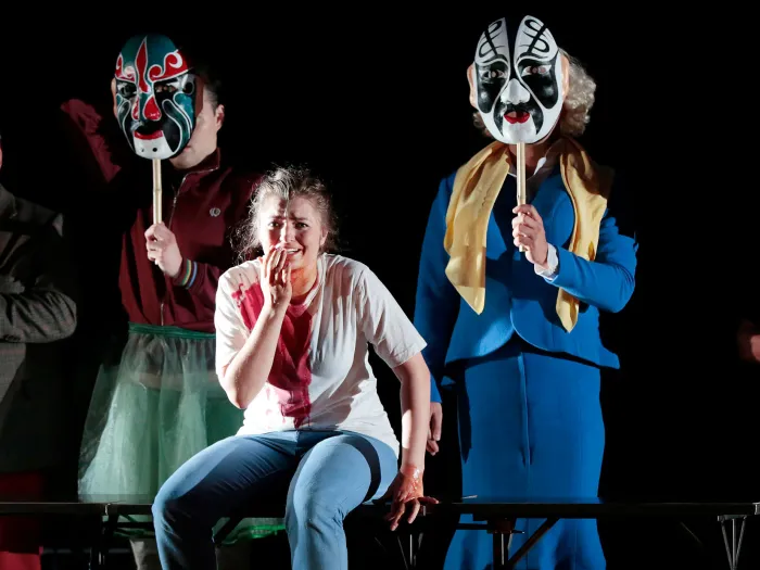 Szene aus "Der goldene Drache", 2015 im Festspielhaus, vier kostümierte Personen halten je eine bunte Maske vor das Gesicht, zentral hockt eine Person und hält ihre linke Hand vor den Mund 
