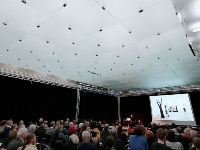 Szene aus "Einblick 2 – Gemeinsam zum Leuchtturm", 2015 im Kunsthaus Bregenz, Publikum sitzt auf Stühlen und blickt nach vorne auf eine Präsentation