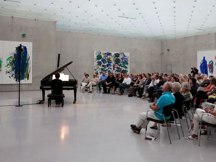 Szene aus "Konzert im KUB", 2015 im Kunsthaus Bregenz, Publikum sitzt auf Stühlen und blickt auf den Pianisten am Flügel, an den Wänden befinden sich großflächige Gemälde