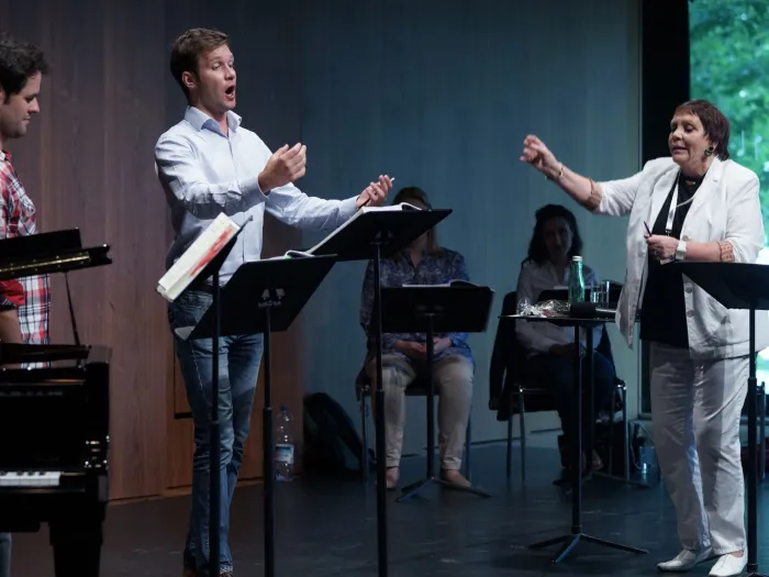 Szene aus "Meisterklasse Brigitte Fassbaender", 2015 im Festspielhaus, rechts Brigitte Fassbaender mit erhobenem Arm, drei weitere Personen stehen hinter einem Pult, zwei weitere Personen sitzen im Hintergrund hinter einem Pult