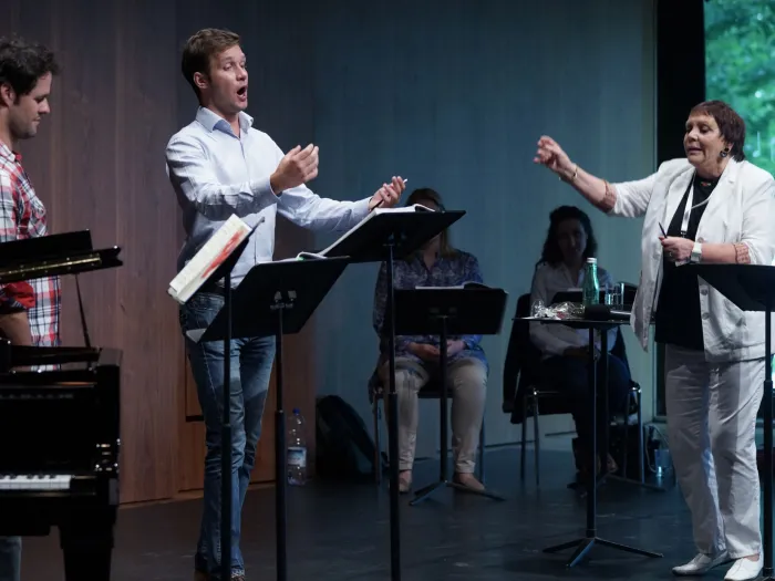 Szene aus "Meisterklasse Brigitte Fassbaender", 2015 im Festspielhaus, rechts Brigitte Fassbaender mit erhobenem Arm, drei weitere Personen stehen hinter einem Pult, zwei weitere Personen sitzen im Hintergrund hinter einem Pult