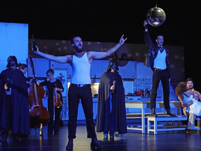 Szene aus "Don Giovanni", 2016 im Theater am Kornmarkt, mehrere kostümierte Darstellende, zentral eine Person in weißem Tanktop mit ausgestreckten Armen, im Hintergrund drei Personen mit Darth-Vader-Maske und schwarzem Umhang, zwei Personen, eine mit einem Kontrabass, die andere mit Geige, rechts steht eine Person auf einer Bank und hält eine Diskokugel, zwei Personen sitzen auf einem Stuhl