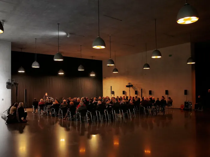 Szene aus "Einblick 3 - Gemeinsam zum Leuchtturm", 2016 im Kunsthaus Bregenz, Publikum sitzt in einem dunklen Raum, mehrere Lampen hängen von der Decke, vor dem Publikum befindet sich eine Person
