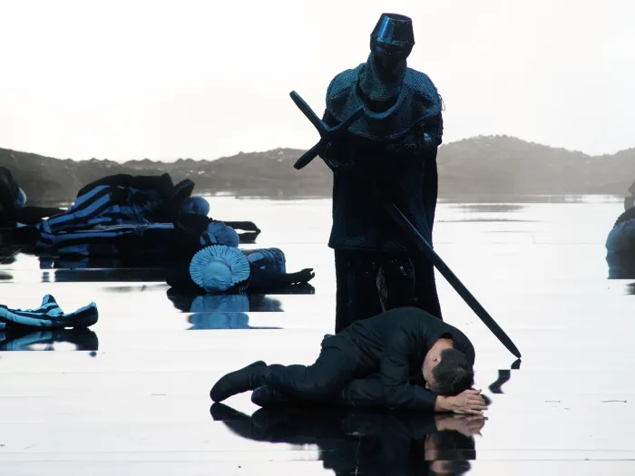 Szene aus "Hamlet", 2016 im Festspielhaus, Ritter in Rüstung mit langem Schwert, davor ein Mann am Boden liegend, reflektierender Boden, im Hintergrund weitere Personen am Boden liegend