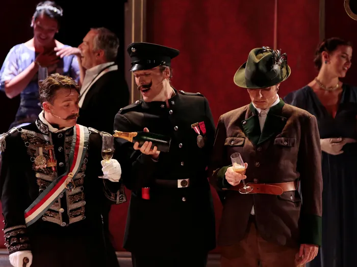 Szene aus "Staatsoperette", 2016 im Festspielhaus, im Vordergrund drei Darsteller in historischen Kostümen, zwei halten Sektgläser, im Hintergrund weitere kostümierte Personen