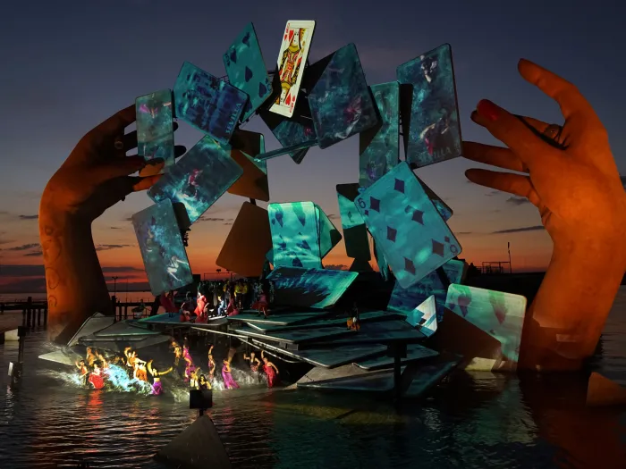 Szene aus "Carmen", 2018 auf der Seebühne, große Hände über dem Wasser, schwebende grün-beleuchtete Spielkarten im Bogen, kostümierte Darstellende im Vordergrund auf der Bühnenfläche