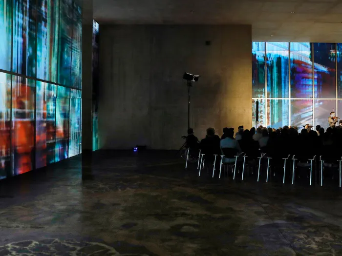 Szene aus "Konzert im KUB", 2017 im Kunsthaus Bregenz, Musiker:in auf der Bühne, davor Publikum auf Stühlen sitzend, Publikum von hinten im Weitwinkel fotografiert