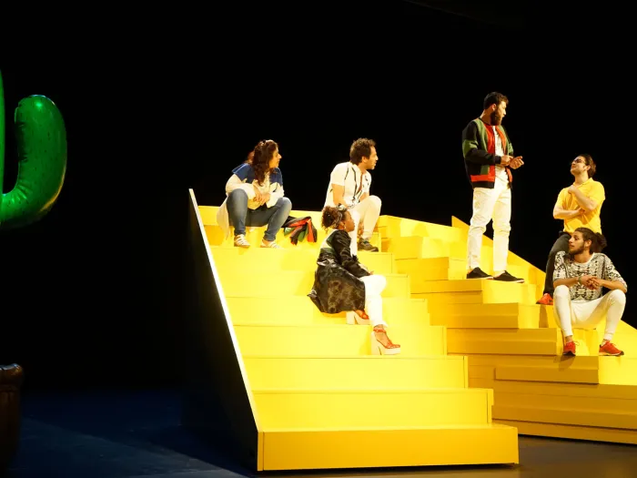 Szene aus "The Situation", 2017 im Theater am Kornmarkt, sechs kostümierte Darstellende auf einer großen gelben Treppe, zwei von ihnen stehen, links ein großer Luftballon in Kaktusform