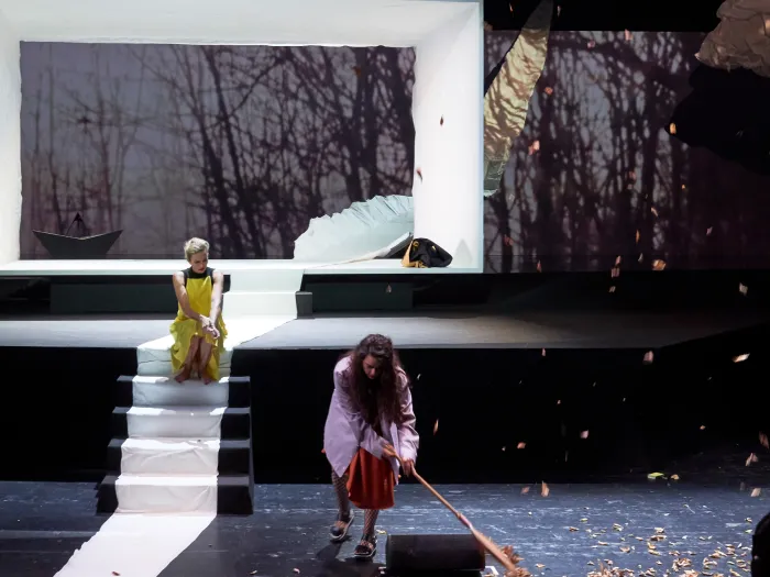 Szene aus "Das Jagdgewehr", 2018 im Festspielhaus, zwei Personen, eine sitzt auf weißen Stufen in gelbem Kleid, die andere kehrt mit Besen auf der Bühne, im im Hintergrund ein übergroßer weißer Rahmen und eine Projektion eines Waldes