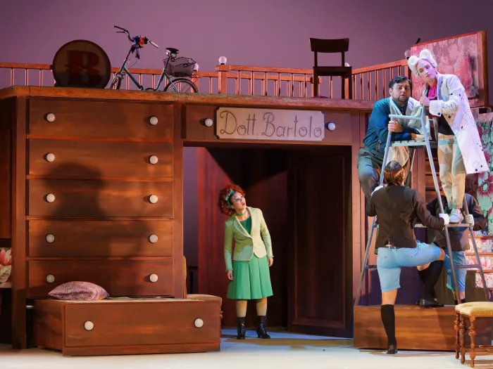 Szene aus "Der Barbier von Sevilla", 2018 im Theater am Kornmarkt, bunt kostümierte Darstellende auf der Bühne, zwei auf einem Hochstuhl, großes Holzmöbelstück mit Schubläden im Hintergrund