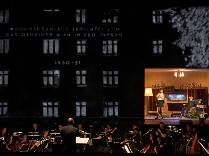 Szene aus "Der Reigen", 2019 im Festspielhaus, Orchester spielt vor Kulisse einer Wohnhausanlage mit Aufschrift "1930-1931", rechts Szene mit zwei kostümierten Darstellenden in Wohnzimmerkulisse
