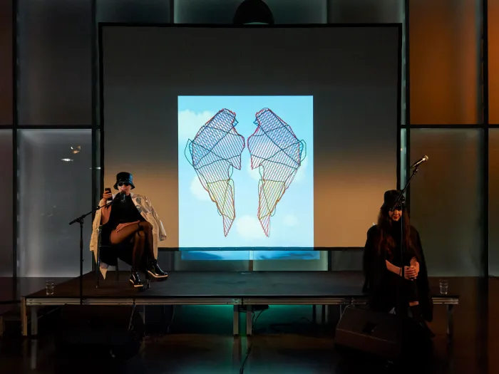 Szene aus "Einblick 2: Die Künstlerin Flaka Haliti", 2019 im Kunsthaus Bregenz, zwei kostümierte Darstellende auf der Bühne, im Hintergrund eine Projektion von zwei stilisierten Flügeln vor blauem Himmel mit Wolken