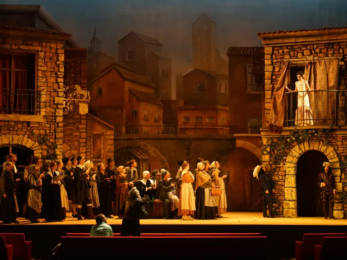 Szene aus "Don Quichotte", 2019 im Festspielhaus, viele kostümierte Darstellende vor Steinhaus-Kulisse, Darstellerin im weißen Kleid auf dem Balkon