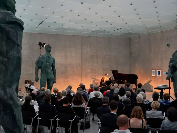 Szene aus "Konzert im KUB", 2019 im Kunsthaus Bregenz, Publikum bestaunt Dame am Klavier, drei übergroße Bronzefiguren umgeben das Publikum