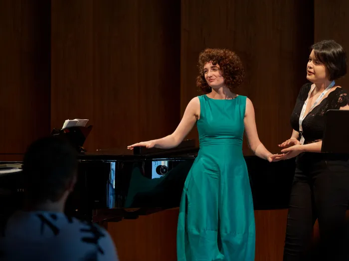 Szene aus "Meisterklasse Dmitry Vdovin", 2019 im Festspielhaus, drei Frauen auf der Bühne, eine im grünem Kleid und hält die Hand der danebenstehenden Frau in schwarzem Kleid, Pianistin am Flügel sitzend