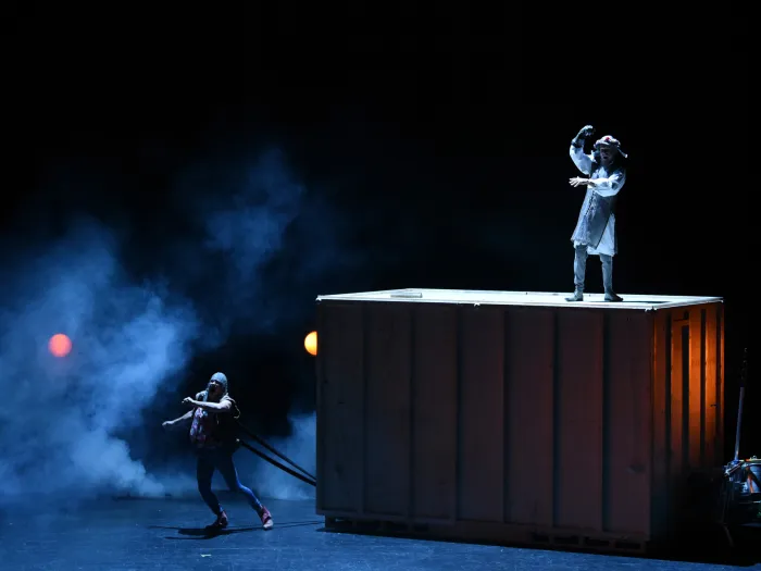 Szene aus "Don Quijote", 2019 im Festspielhaus, kostümierte Darsteller:in steht auf großem Kasten, zwei weitere kostümierte Darsteller unten am Kasten in ziehender Position befestigt