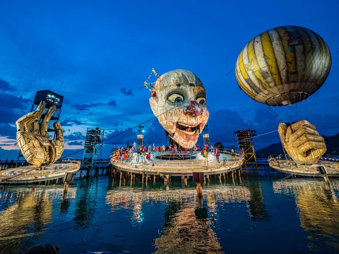 Szene aus "Rigoletto", 2019 auf der Seebühne, riesiger Kopf mit leicht geöffnetem Mund, zwei große Hände, eine Hand hält einen Ballon, mehrere kostümierte Darstellende auf der Bühnenplattform unter dem Kopf