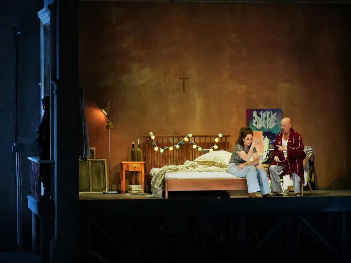 Szene aus "Tancredi", 2024 im Festspielhaus, zwei Personen auf einem Bett, eine Frau und ein Mann, im Hintergrund ein Kreuz an der Wand, Lichterkette über dem Bettende, Nachtkästchen und Lampe links neben dem Bett 