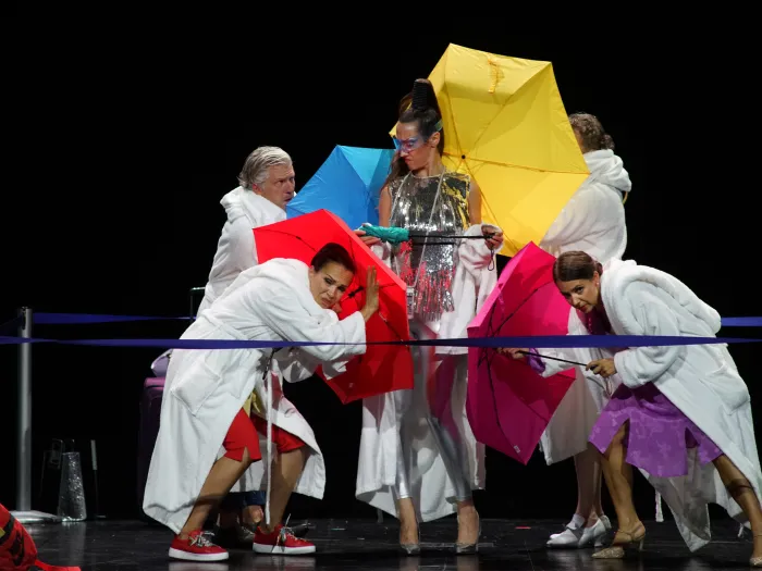 Szene aus "Impresario Dotcom", 2020 im Festspielhaus, fünf Personen, in der Mitte eine Darstellerin in glänzendem Kleid, die anderen tragen weiße Bademantel und halten bunte Regenschirme