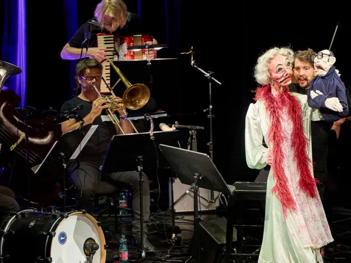 Szene aus "Alles nicht wahr", 2021 im Festspielhaus, Puppe mit weißen Locken und rotem Schal in weißem Kostüm, daneben ein Mann, Musiker:innen im Hintergrund