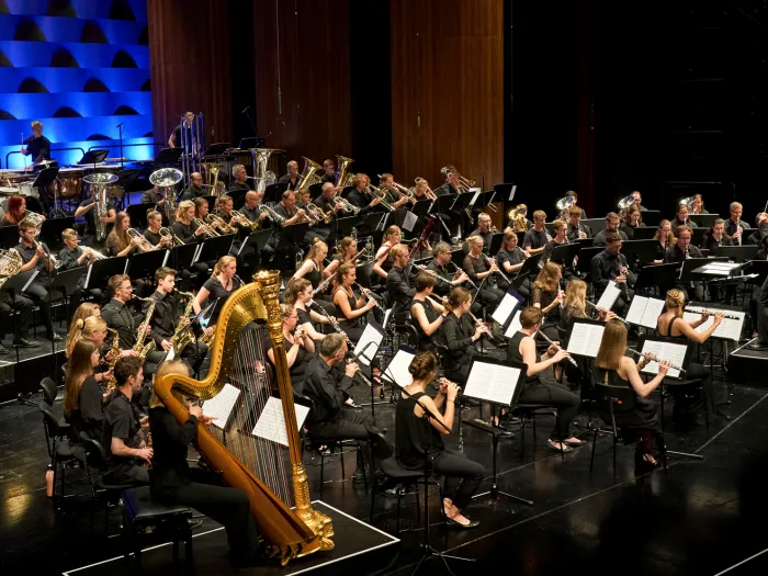 Szene aus "Brass Eroico", 2021 im Festspielhaus, großes spielendes Orchester mit Streichern, Bläsern und einer Harfe, Dirigent davorstehend