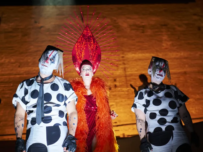 Drei Personen in Bühnenkostümen: zwei in schwarz-weiß gepunkteten Outfits, eine in rotem Kleid mit großem, rotem Kopfschmuck und orange Federboa