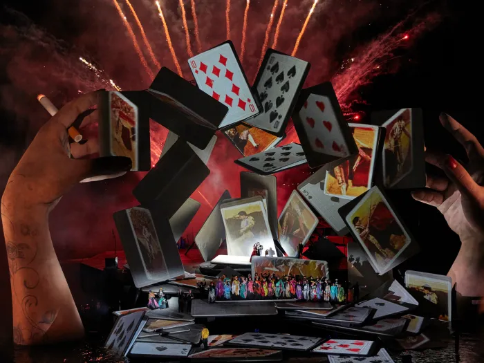 Szene aus "Carmen", 2018 auf der Seebühne, große Hände über dem Wasser, schwebende Spielkarten im Bogen, kostümierte Darstellende im Vordergrund auf der Bühnenfläche, rotes Feuerwerk im Hintergrund