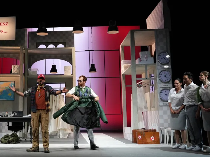 Szene aus "Der Ehevertrag", 2024 im Theater am Kornmarkt, zwei Schauspieler in der Mitte, einer hält eine grüne Jacke, rechts vier Personen in weißer Kleidung, links ein Leuchtschild mit der Aufschrift "Bregenz Brides", weiße Brautkleider auf Kleiderstangen