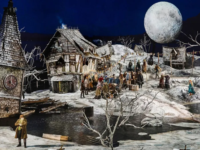 Szene aus "Der Freischütz", 2024 auf der Seebühne, schneebedecktes Dorf, links ein Turm mit Uhr, im Hintergrund ein großer Mond, viele kostümierte Darstellende auf der Bühne verteilt