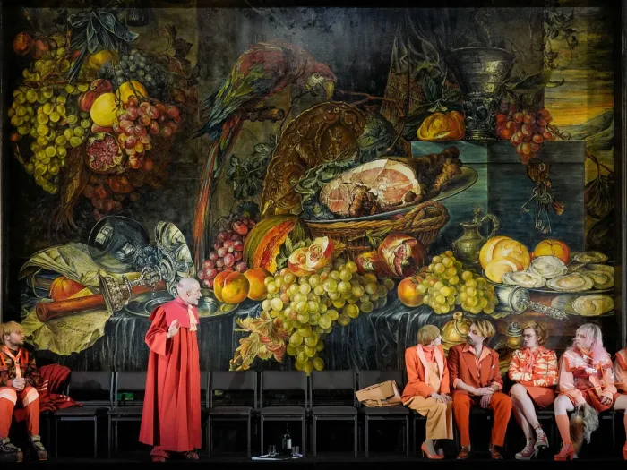 Szene aus "Der zerbrochene Krug", 2024 im Theater am Kornmarkt, sieben Darstellende in rot-orangen Kostümen vor einem großen Stillleben-Gemälde mit Früchten, Fleisch und einem Papagei
