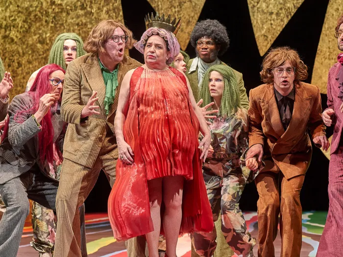 Szene aus "Die gefesselte Phantasie", 2023 im Festspielhaus, elf kostümierte Darstellende, zentral eine Person in rotem Kleid und mit Krone auf dem Kopf