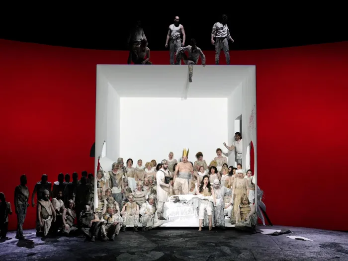 Szene aus "Ernani", 2023 im Festspielhaus, viele weiß kostümierte Darstellende in großer Kubusstruktur, roter Hintergrund, links weitere Darstellende im Schatten