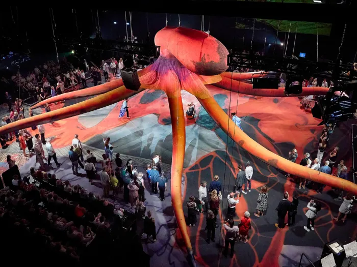 Szene aus "Hold Your Breath", 2024 im Festspielhaus, großer, roter Oktopus in der Mitte, umgeben von Zuschauende 