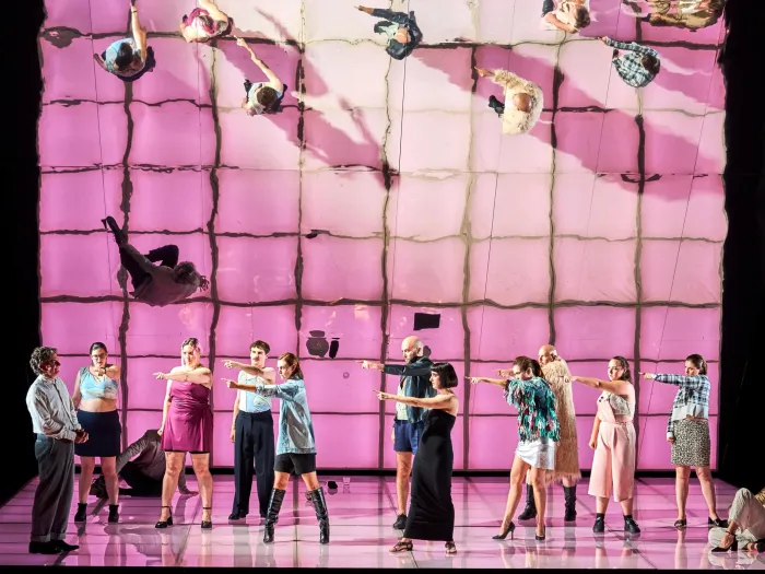 Szene aus "Die Judith von Shimoda", 2023 im Festspielhaus, zwölf kostümierte Darstellende, elf auf eine Person zeigend, rosa-farbender Boden, spiegelnde Wand im Hintergrund