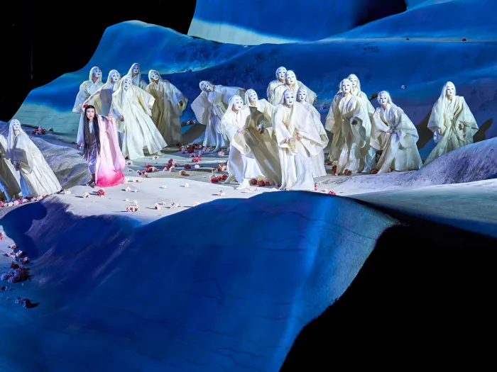 Szene aus "Madame Butterfly", 2022 auf der Seebühne, wellenförmige Bühne, rosafarbene Blüten auf dem Boden verteilt, viele Darstellende in weißen, langen Gewändern mit weißen Gesichtsmasken, links im Bild die Darstellende Madame Butterfly mit langem, dunklen Haar und rosafarbenem Gewand