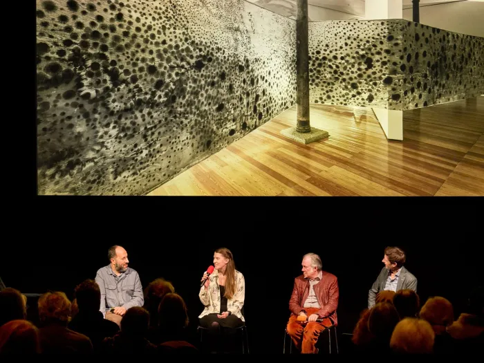 Szene aus "Einblick 2", 2023 im Festspielhaus, vier Personen auf der Bühne vor einem Publikum, im Hintergrund ein Bild eines großformatigen Kunstwerks mit schwarzen Punkten auf hellem Untergrund