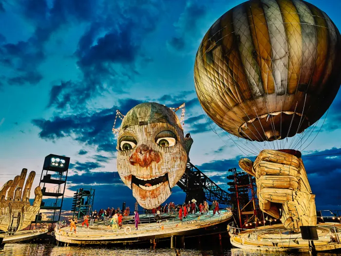 Szene aus "Rigoletto", 2021 auf der Seebühne, riesiger Kopf mit leicht geöffnetem Mund, zwei große Hände, eine Hand hält einen Ballon, mehrere kostümierte Darstellende auf der Bühnenplattform unter dem Kopf