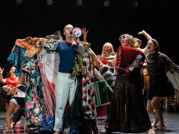 Szene aus "The Faggots and Their Friends Between Revolutions", 2023 im Festspielhaus, acht singende und kostümierte Darstellende, eine Person mit Megafon in der Hand, eine weitere mit einem Akkordeon
