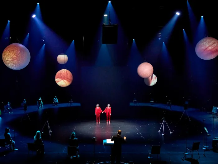 Szene aus "Unmögliche Verbindung", 2024 im Festspielhaus, von der Decke hängende Planetenmodelle, in der Mitte zwei sich an Händen haltende Darstellende in rotem Kostüm