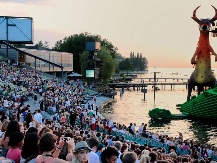 Szene aus "crossculture", 2014 an diversen Spielorten, viele Menschen sitzen auf der Tribüne, rechts ein Teil des Bühnenbilds von "Die Zauberflöte", im Hintergrund der Sonnenuntergang