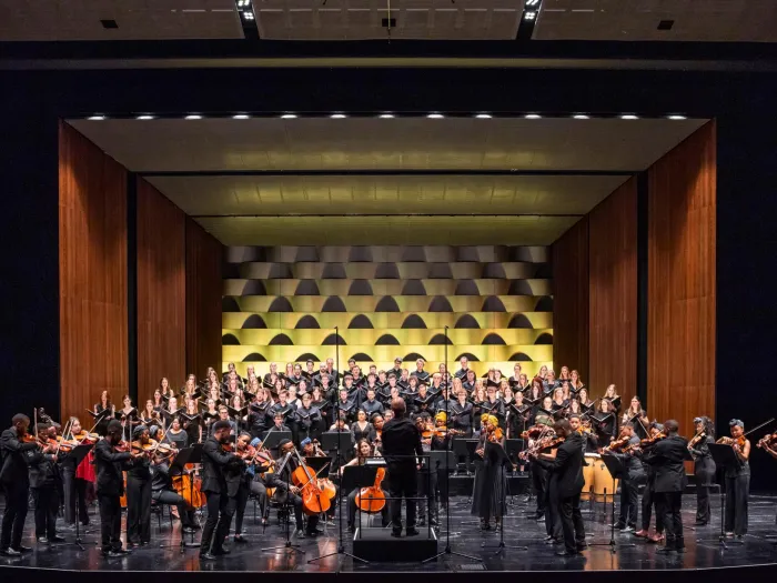 Szene aus "Zwischen Himmel und Erde", 2023 im Festspielhaus, Orchester stehend und spielend im Vordergrund auf der Bühne, Chor im Hintergrund
