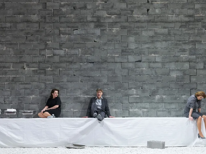Szene aus "Geschlossene Gesellschaft", 2022 im Festspielhaus, drei kostümierte Darstellende, sitzend auf einer Tafel, Tisch mit weißer Tischdecke bis zum Boden bedeckt, graue Steinmauer als Hintergrund 